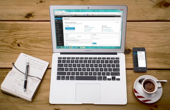 Wooden desk with coffee, notebook, phone and laptop with back end of website displayed on the screen that benefits from cheap WordPress hosting.